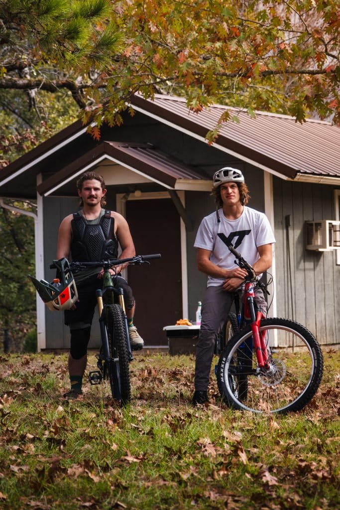 Two Hustle Bike Labs riders sitting on their bikes in front of a shed with fall foliage all around them.